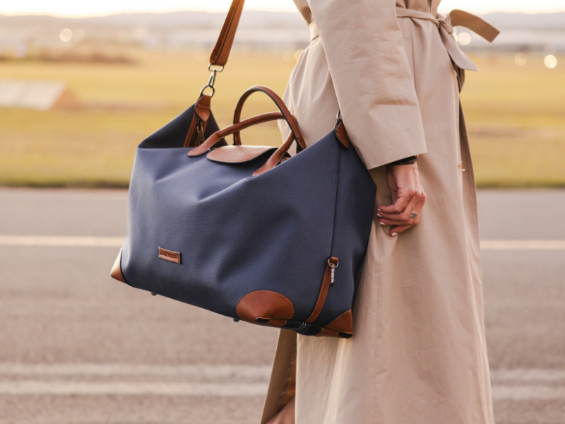 A woman wearing a trench coat and carrying a navy nylon overnight bag on her shoulder