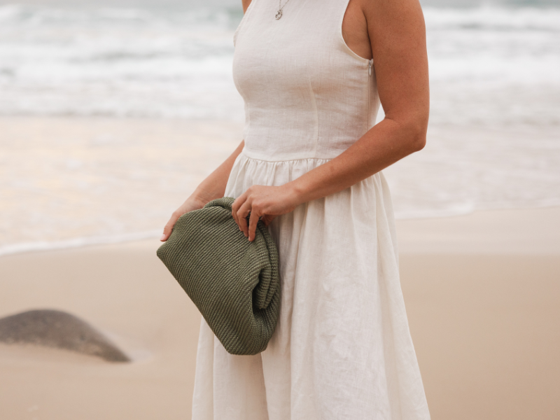 A woman wearing a cream linen dress holding a sage green, raffia dumpling clutch