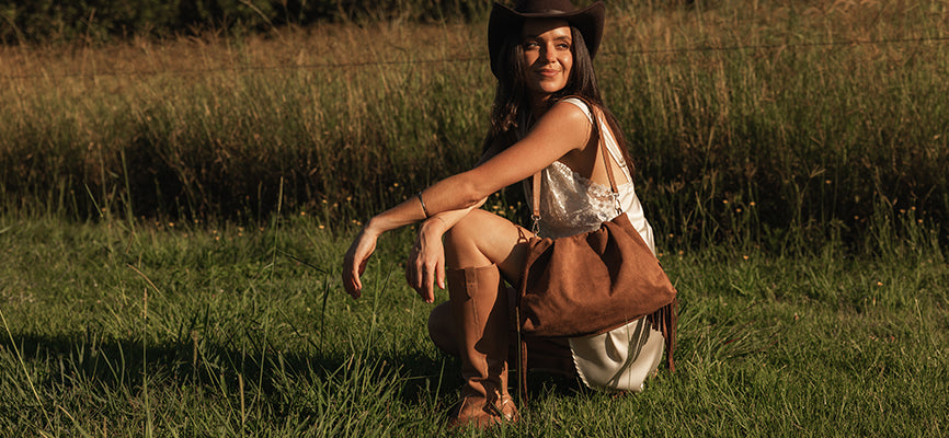 Louenhide Reese Suedette Shoulder Bag hangs from womans shoulder as she kneels in field