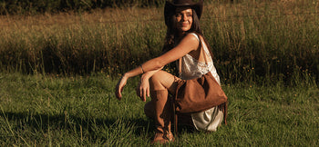 Louenhide Reese Suedette Shoulder Bag hangs from womans shoulder as she kneels in field