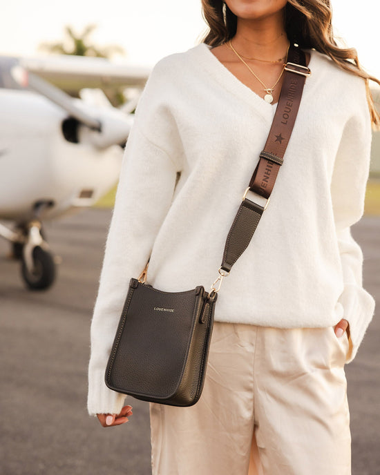 Louenhide Parker Chocolate Phone Bag is worn crossbody over white sweater and pants in airfield