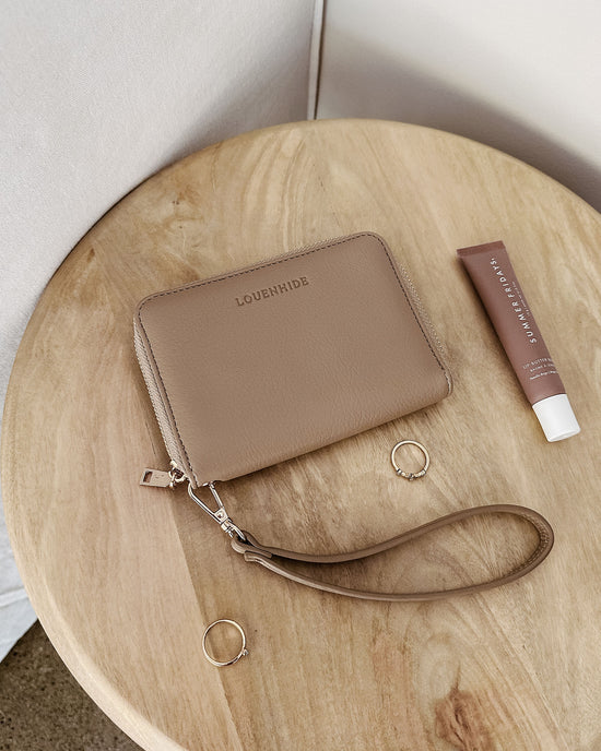 The Eden Wallet - Malt by Louenhide, a Summer Fridays lip balm, and two gold rings are arranged on a round wooden table against a neutral background.